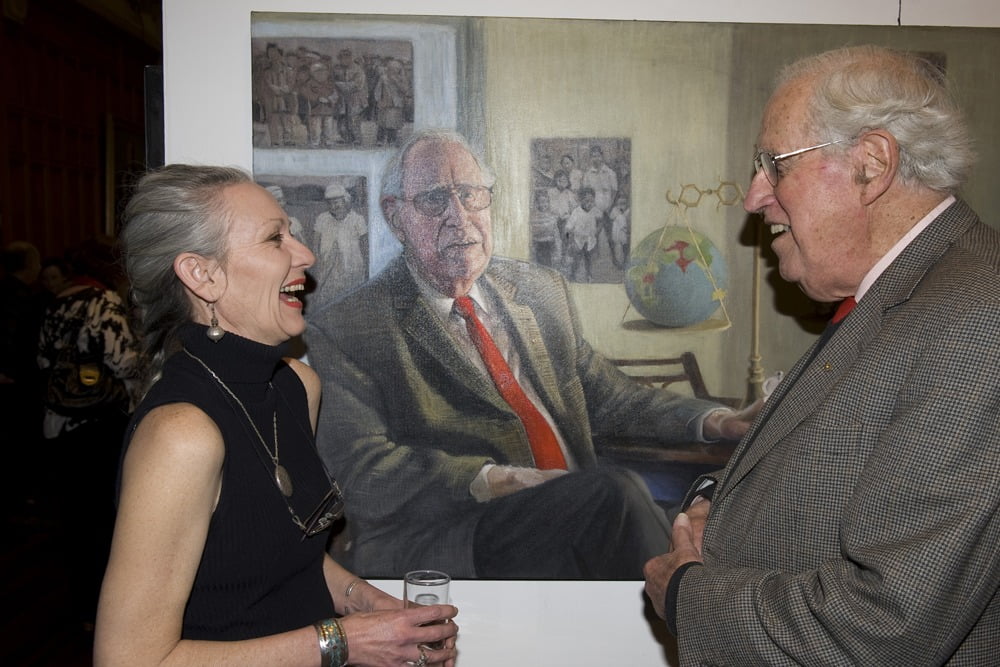 Avril Thomas and Basil Hetzel smiling and chatting at the University of Adelaide School of Medicine exhibition opening, standing proudly in front of Thomas's painted portrait of Hetzel, which celebrates his groundbreaking work discovering the link between iodine deficiency and brain damage, leading to global efforts to prevent iodine deficiency disorders.