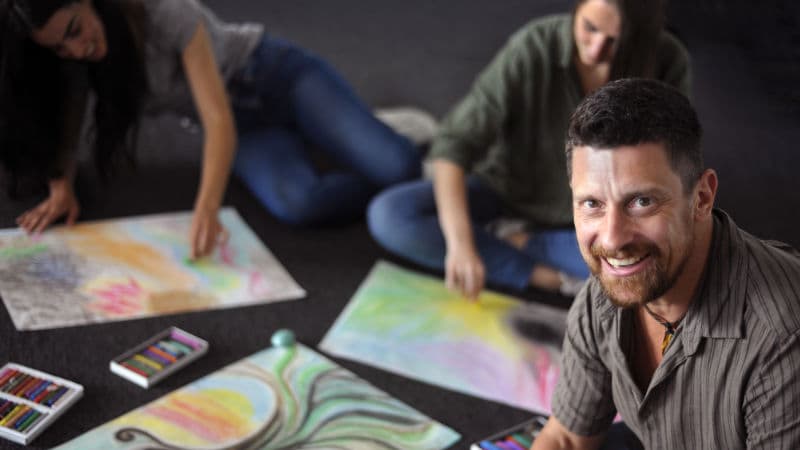 A smiling Advanced Diploma of Transpersonal Therapy instructor with students artwork.