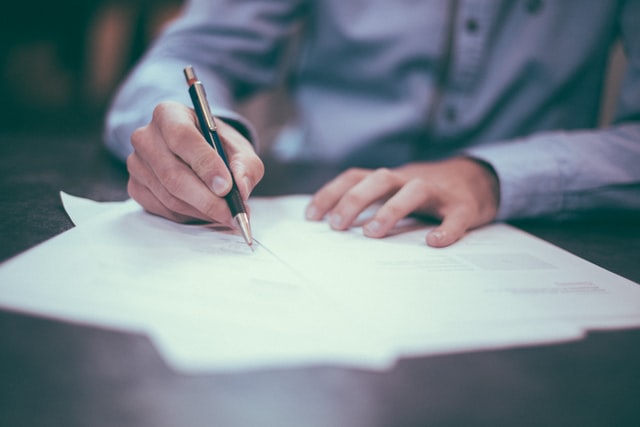 Person in a blue shirt signing documents with a pen on a desk