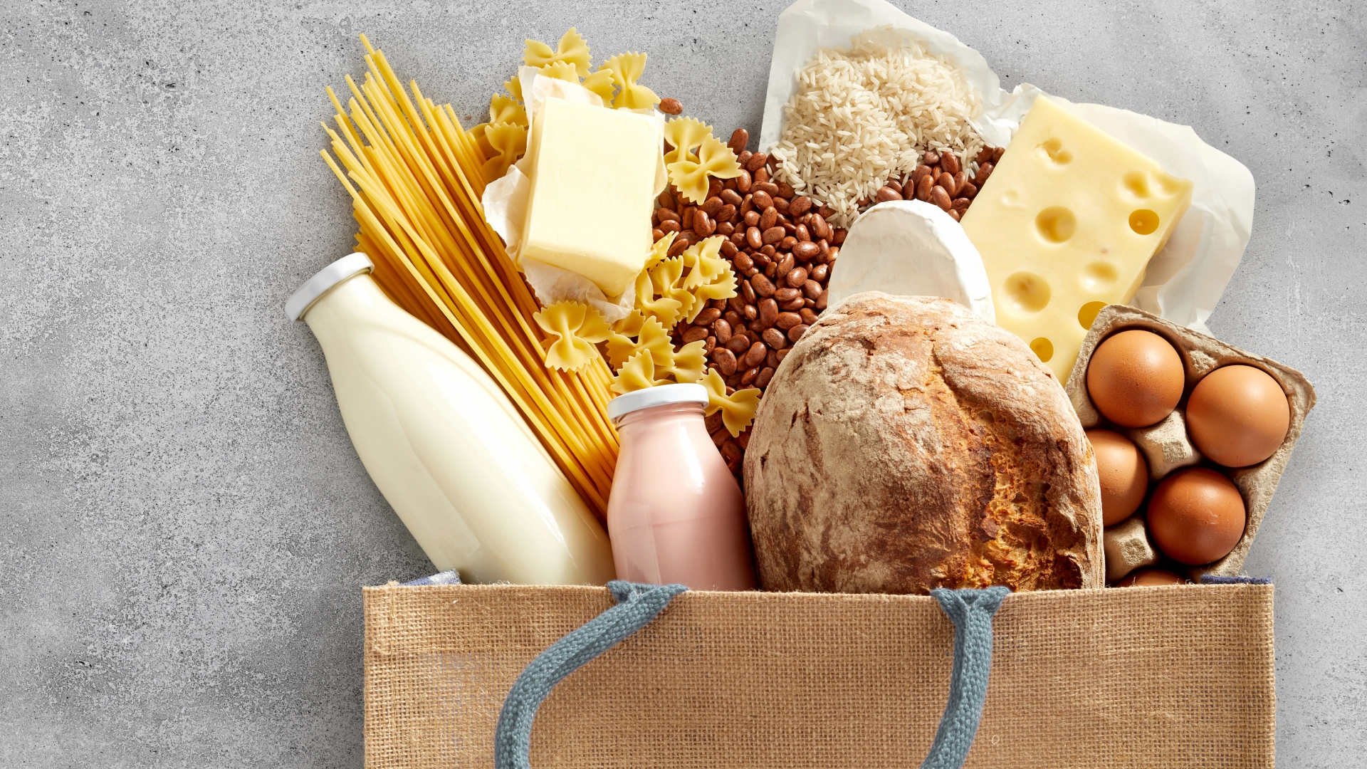 A shopping bag bursting with foods including milk, pasta, fresh bread, eggs, cheese and rice
