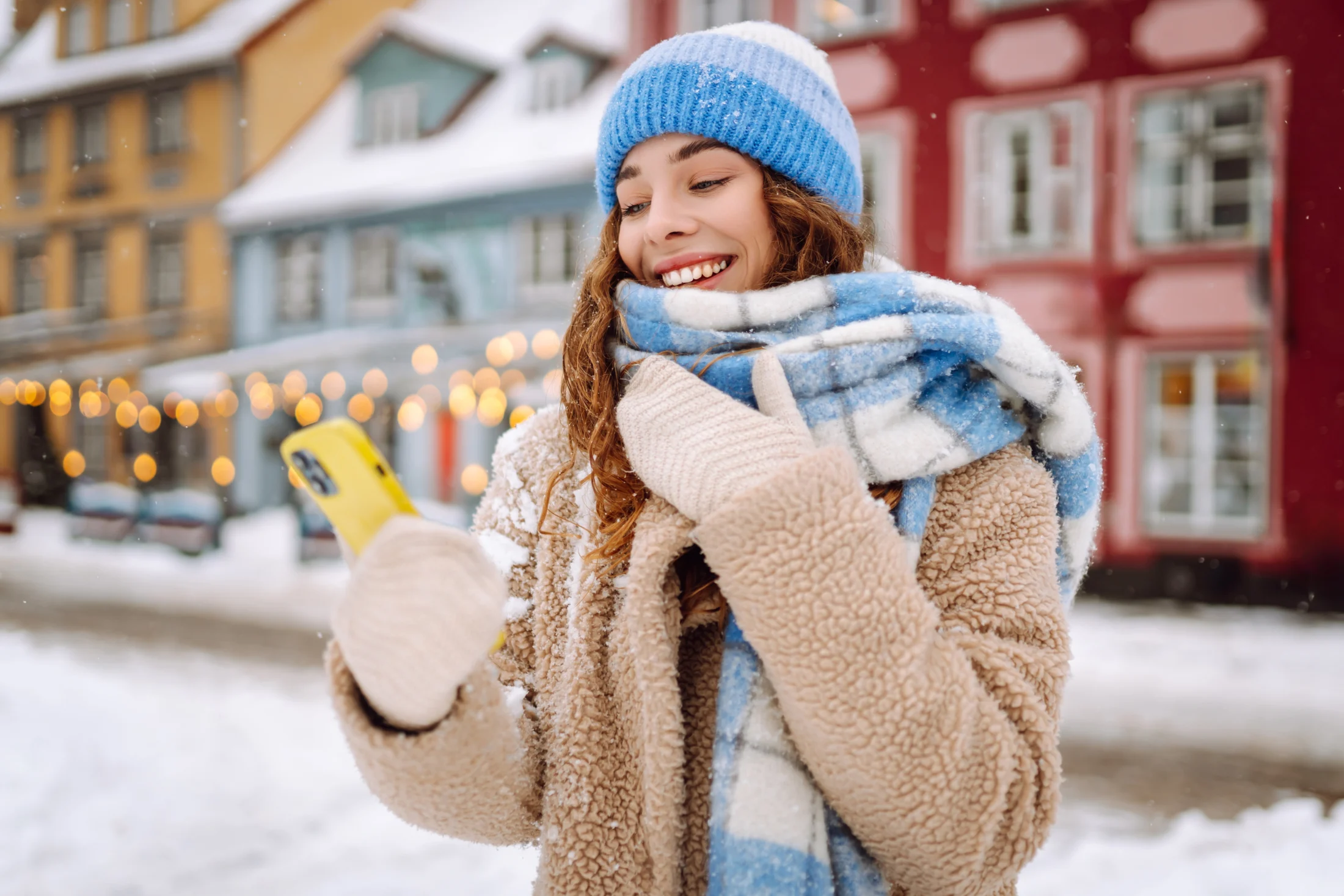 A woman bundled in winter clothing smiles at her phone in a snowy, festive town, appearing to check or apply for a holiday loan with Swoosh Finance.