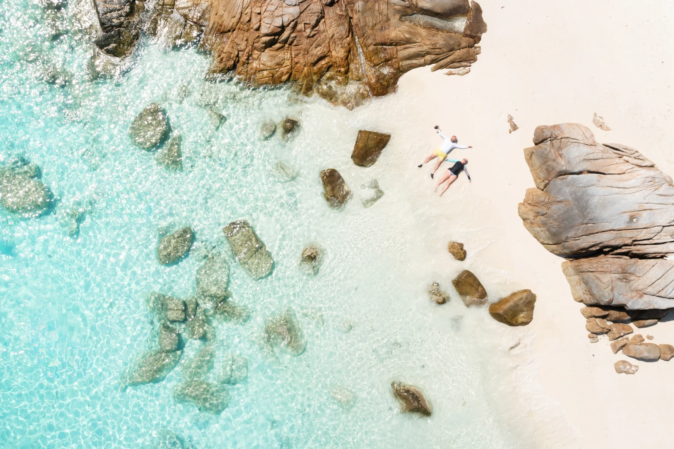 An aerial view of two people relaxing on a white-sand beach beside clear turquoise water and large coastal rocks.